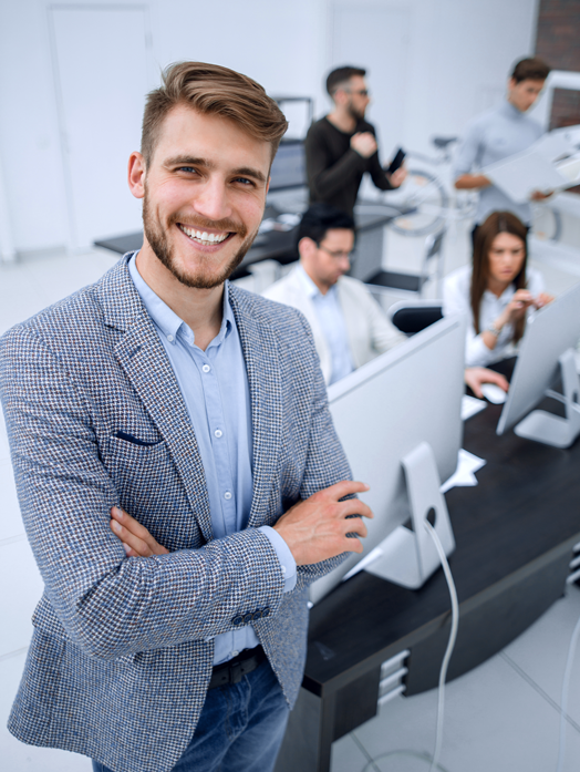 Mann steht mit verschränkten Armen vor seinen KollegInnen, die an PCs arbeiten und lächelt in die Kamera